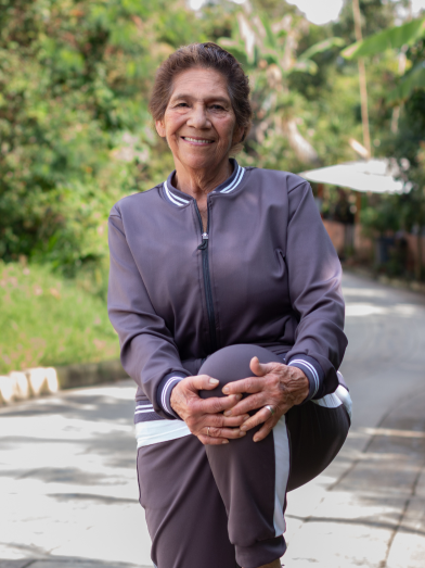 An older woman standing on one leg outside and stretching.